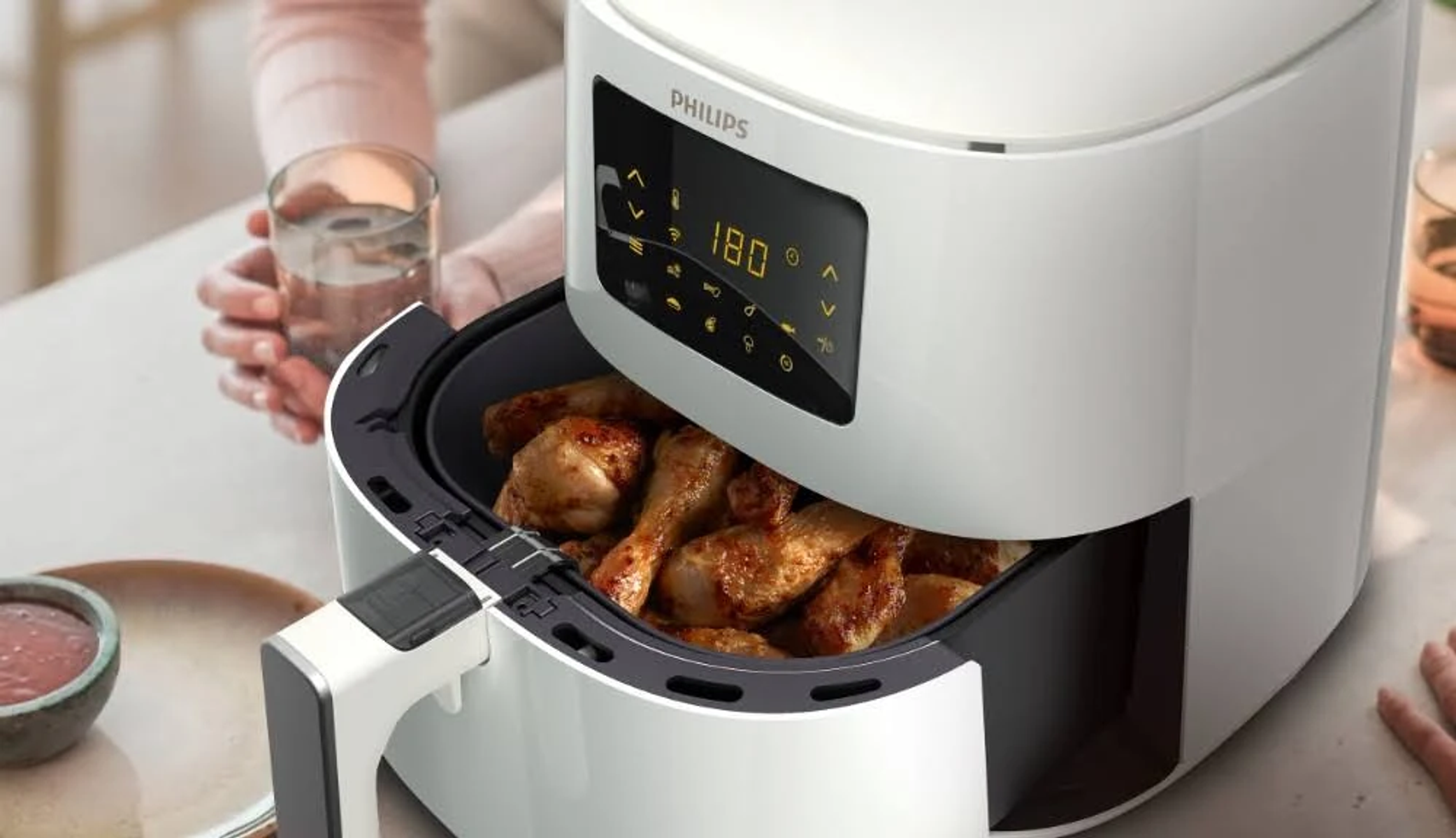 Biała frytkownica beztłuszczowa z jedzeniem w środku, dłonią w pobliżu i stołem z innym jedzeniem.