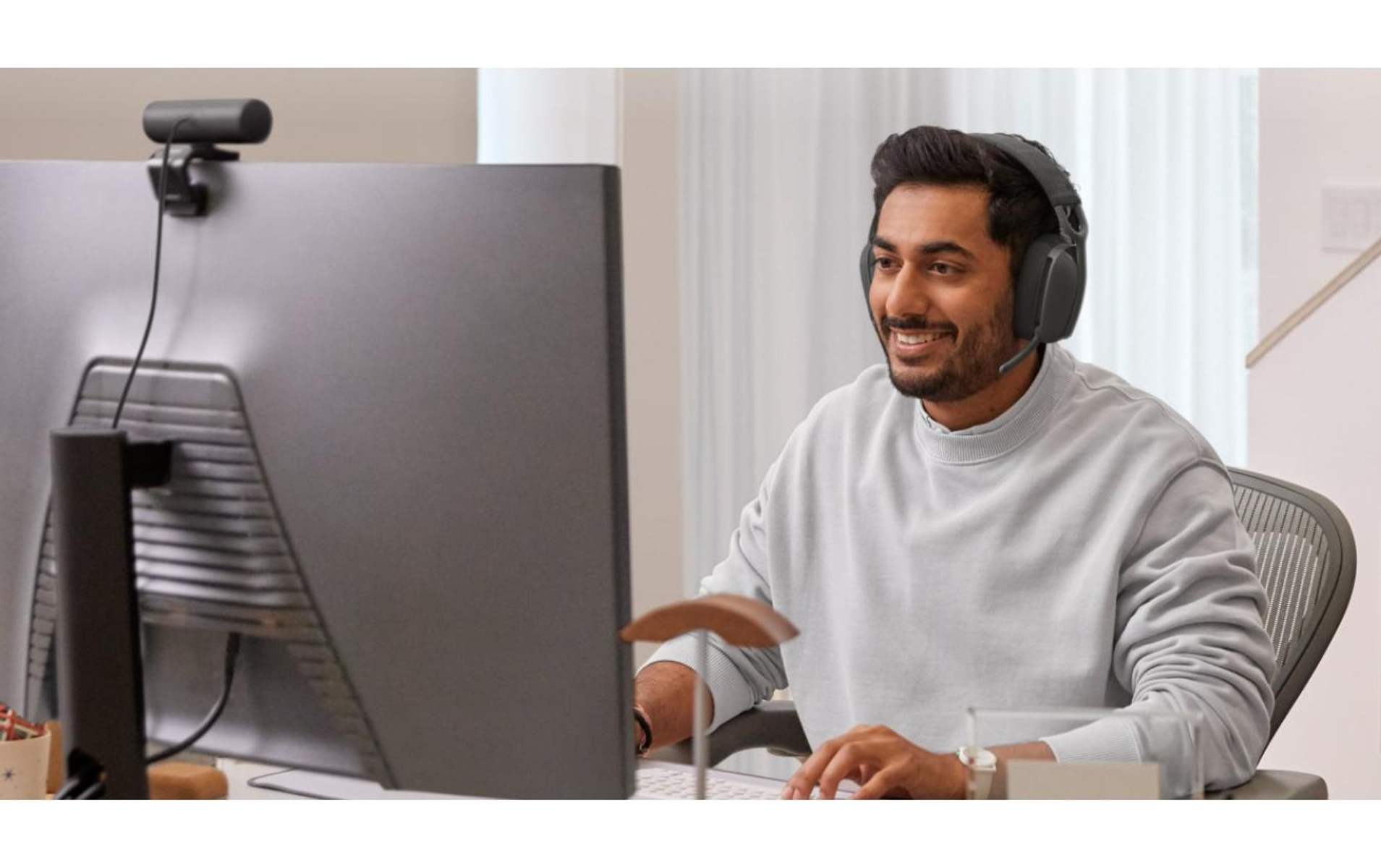 Mężczyzna siedzi przed komputerem z zestawem słuchawkowym i kamerą internetową, uśmiechając się. Tłem jest domowe biuro.