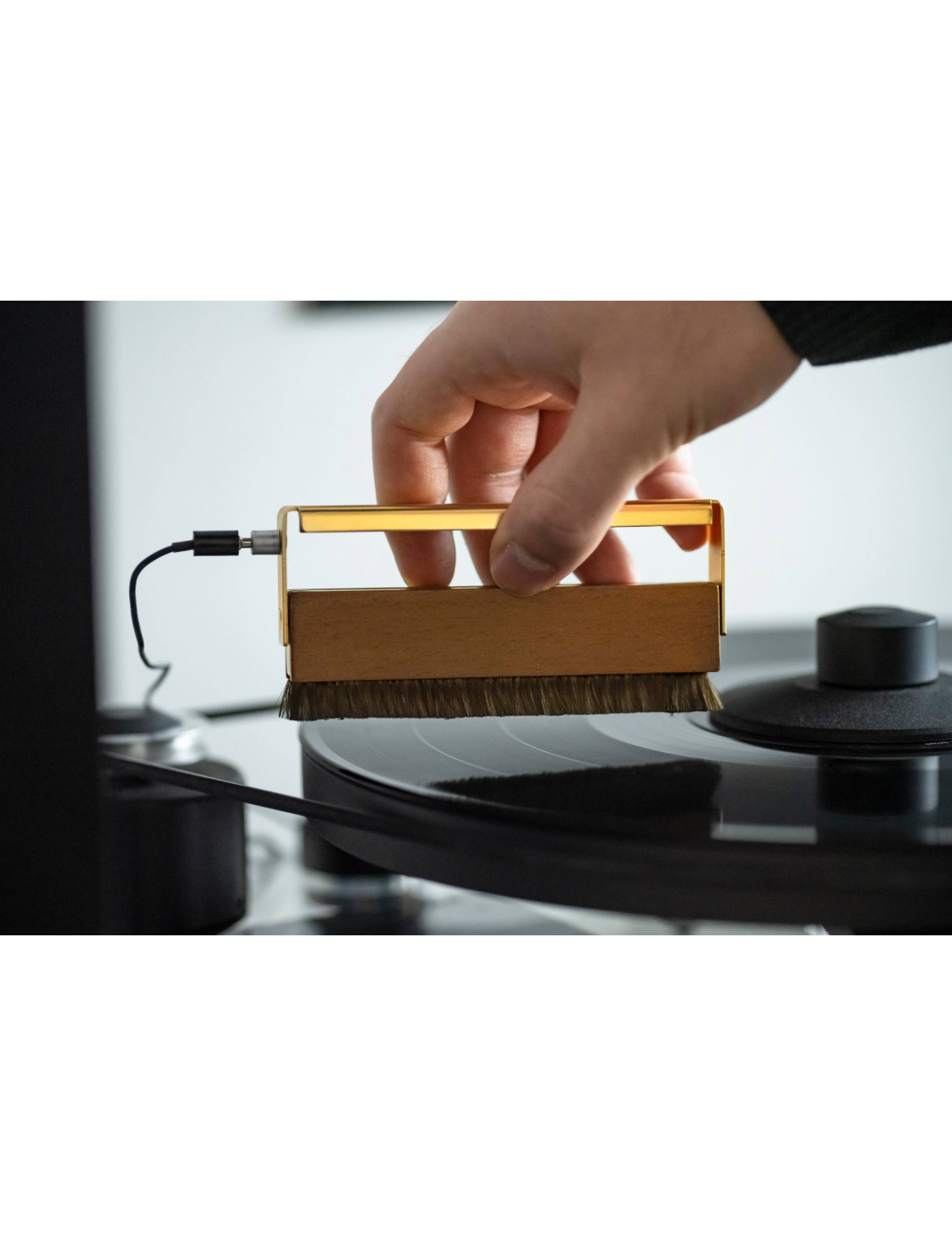 Ręka trzymająca szczotkę do czyszczenia płyt na płycie winylowej; złoty uchwyt szczotki i czarny kabel.