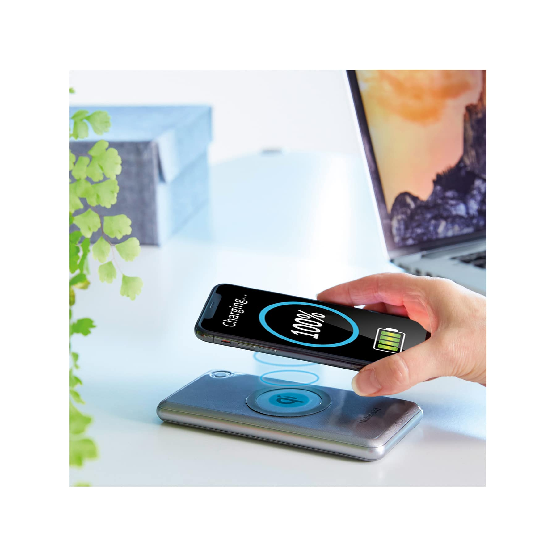 Una mano che carica un telefono su un caricatore wireless; un laptop sullo sfondo.