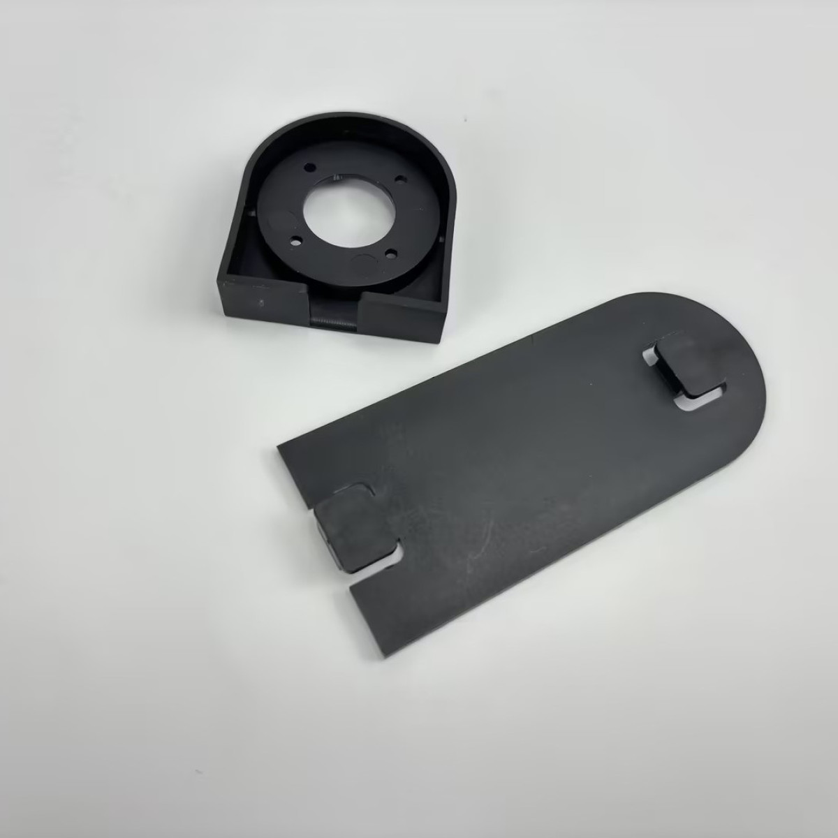 Schwarze Kunststoffteile, eine Halterung und eine Abdeckung, werden auf einer weißen Oberfläche angezeigt.