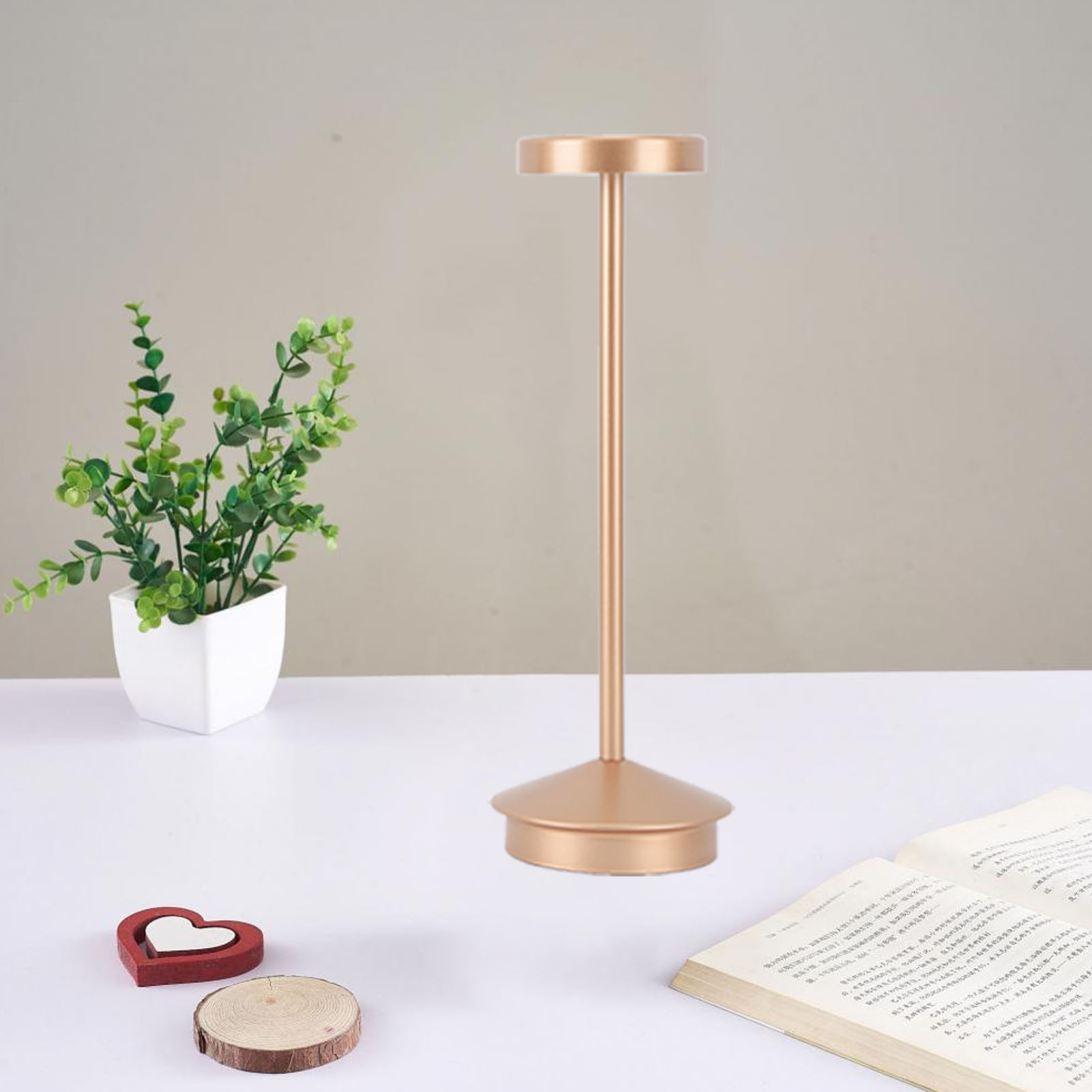 Eine goldene Tischlampe mit schmalem Stiel und kreisförmigem Fuß. Eine Pflanze und ein Buch sind im Hintergrund.
