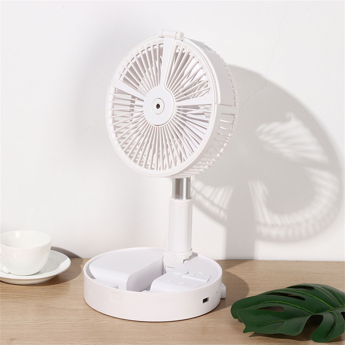 Ein weißer, faltbarer Ventilator auf einem Holztisch, mit einem Blatt und einer Teetasse daneben.