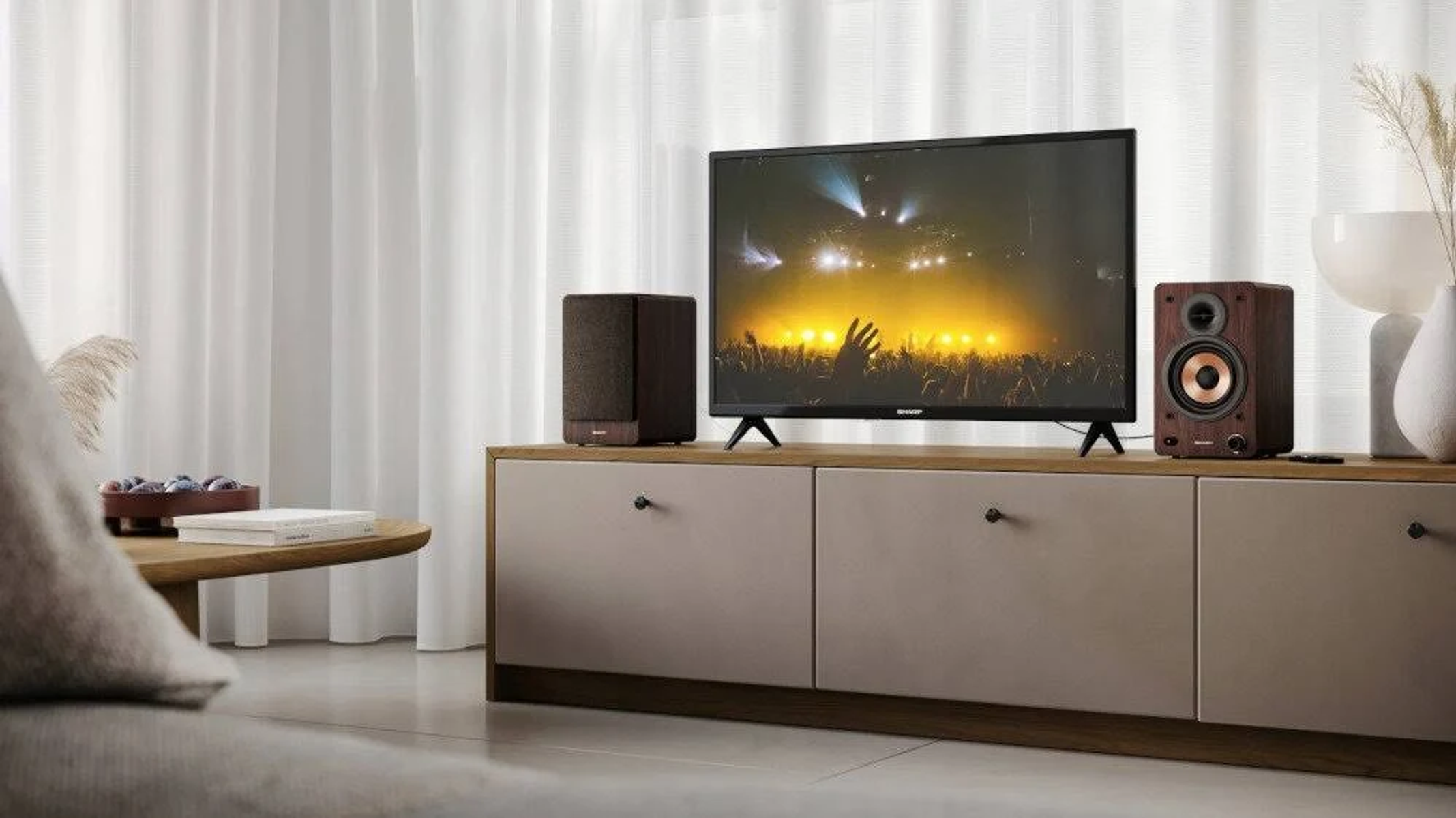 Televisor con altavoces en un mueble. El televisor muestra un concierto. Fondo con cortinas y sofá.