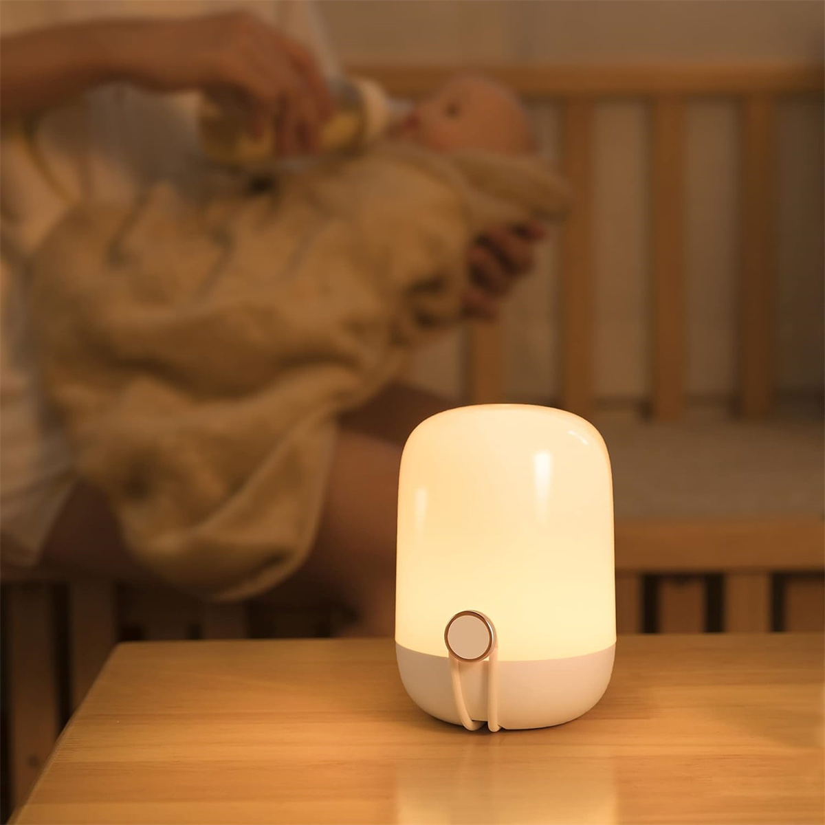 Eine beleuchtete, weiße und beige Nachttischlampe auf einem Holztisch. Im Hintergrund füttert eine Person ein Baby.