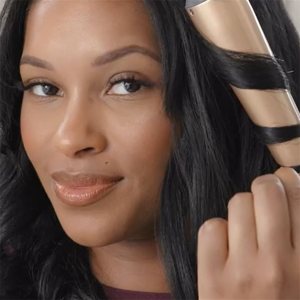 Frau lockt ihr Haar mit einem Lockenstab. Sie lächelt, mit dunklem Haar und Make-up.