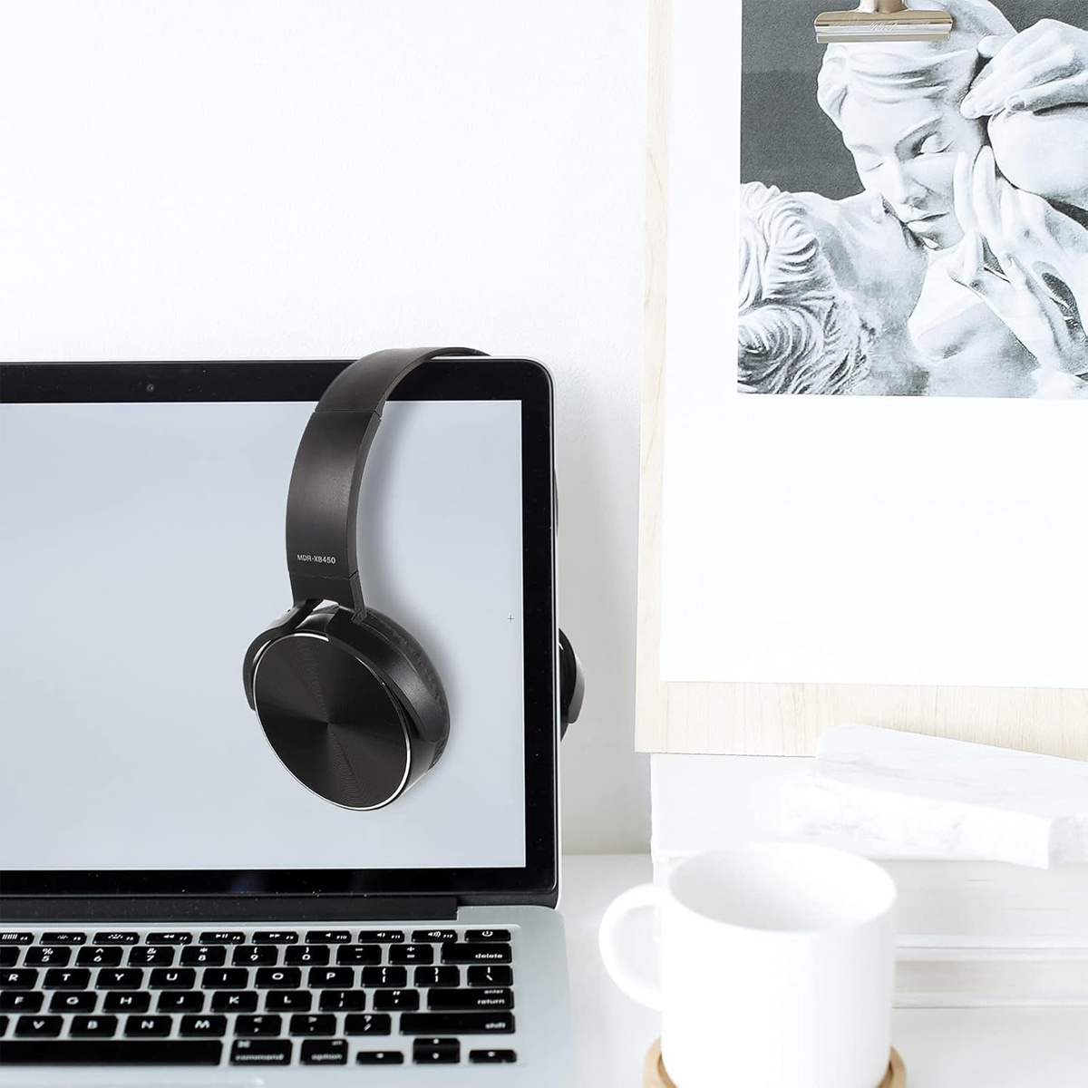 Laptop mit Kopfhörern, die am Bildschirm hängen. Daneben eine weiße Tasse und ein Bild.