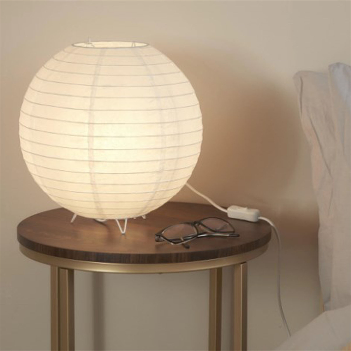Runde Papierlampe auf einem Holztisch mit Brille. Der Hintergrund ist eine hellfarbige Wand und Bett.