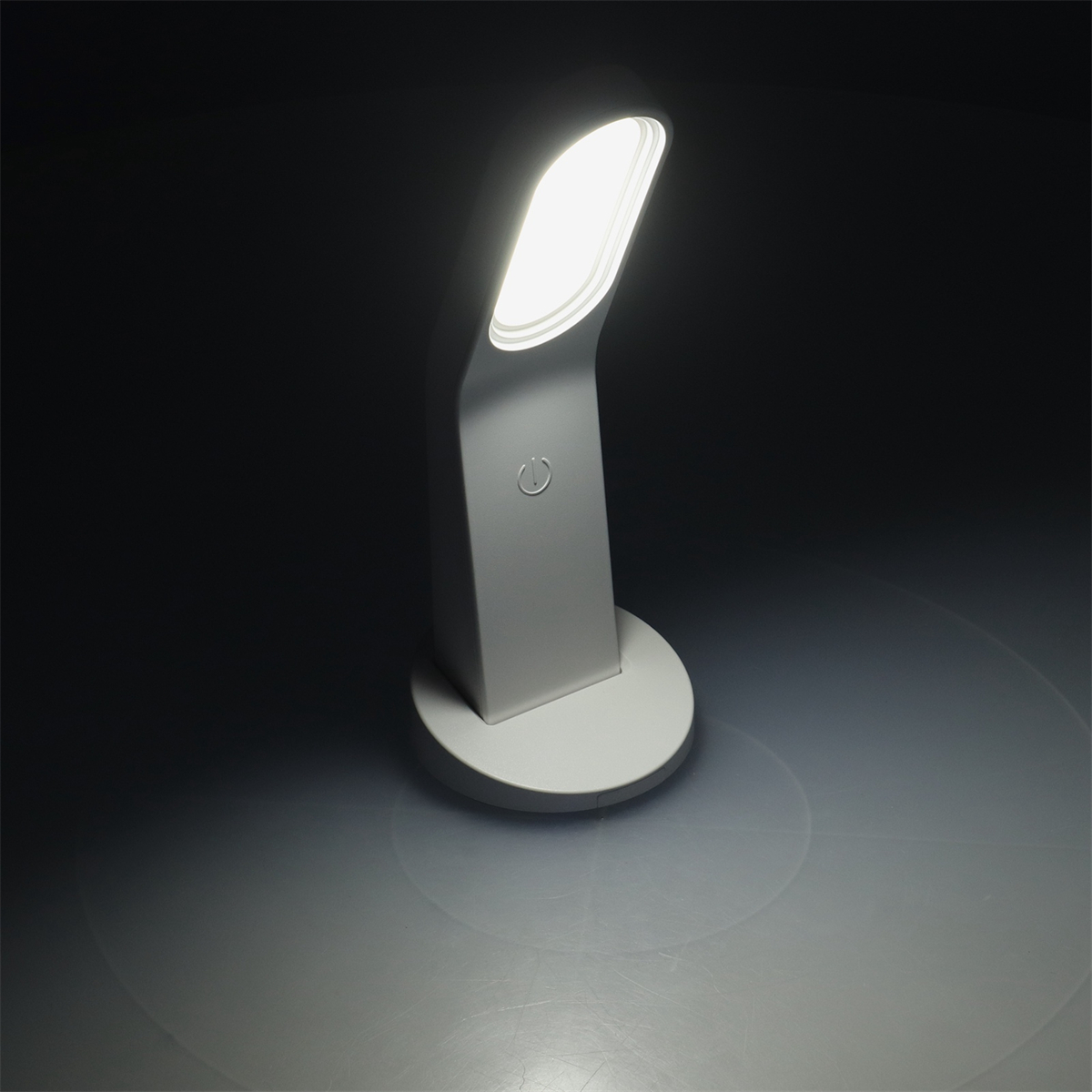 Weiße Schreibtischlampe, die helles Licht ausstrahlt. Die Lampe steht auf weißem Sockel, dunkler Hintergrund.