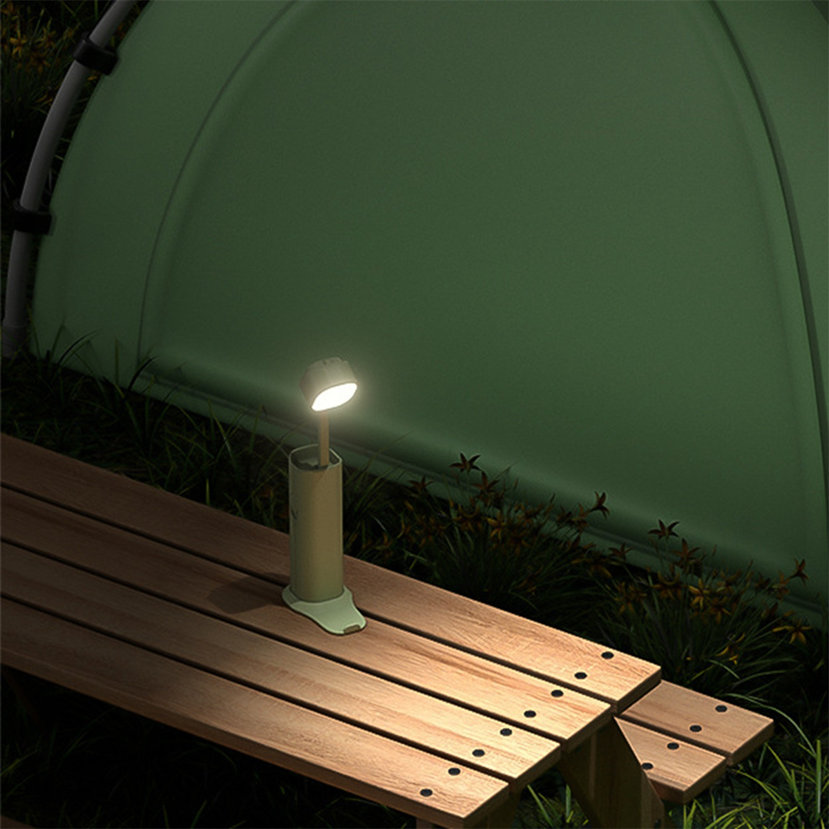 Eine beleuchtete grüne Campinglampe steht auf einem Picknicktisch mit Zelt und Gras im Hintergrund.
