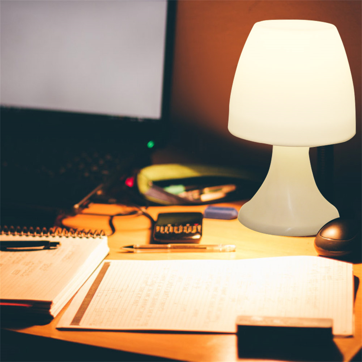 Eine weiße pilzförmige Lampe auf einem Schreibtisch. Notizbuch, Stift und Computer im Hintergrund.