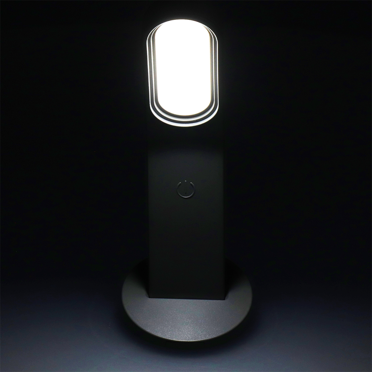 Eine schwarze Lampe mit einer hellen, ovalen Lichtquelle. Der Sockel ist kreisförmig und ruht auf einer Oberfläche.