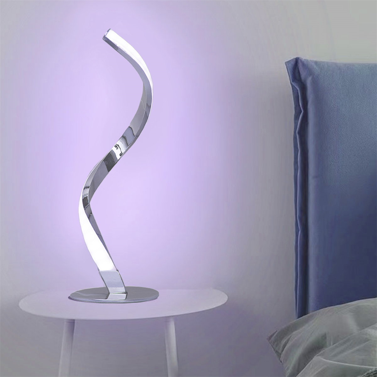 Eine silberne, wellenförmige Schreibtischlampe auf einem weißen Tisch. Bett und Wand im Hintergrund.