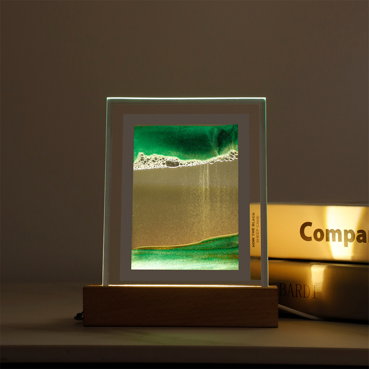 Beleuchtete Sandkunst in einem Glasrahmen auf Holzsockel, dahinter ein Buch.
