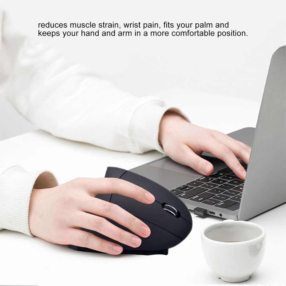 Die Hände einer Person, die eine schwarze vertikale Maus halten, mit einem Laptop und einer Tasse in der Nähe.