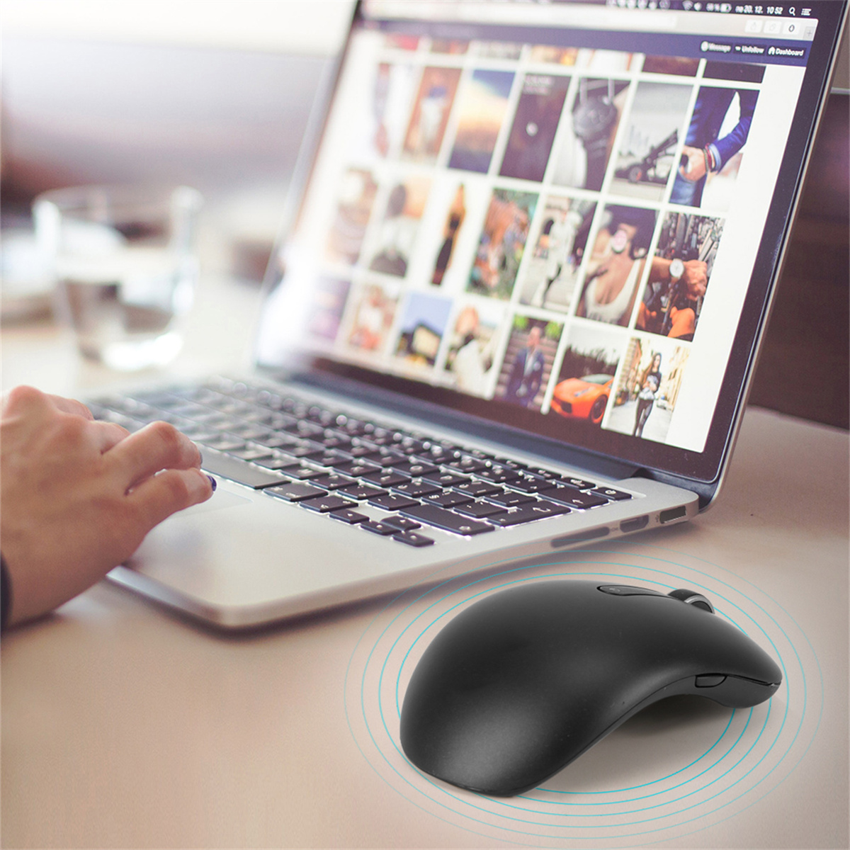 Schwarze Maus auf einem Schreibtisch mit Laptop im Hintergrund. Ein Glas Wasser und eine Hand sind sichtbar.