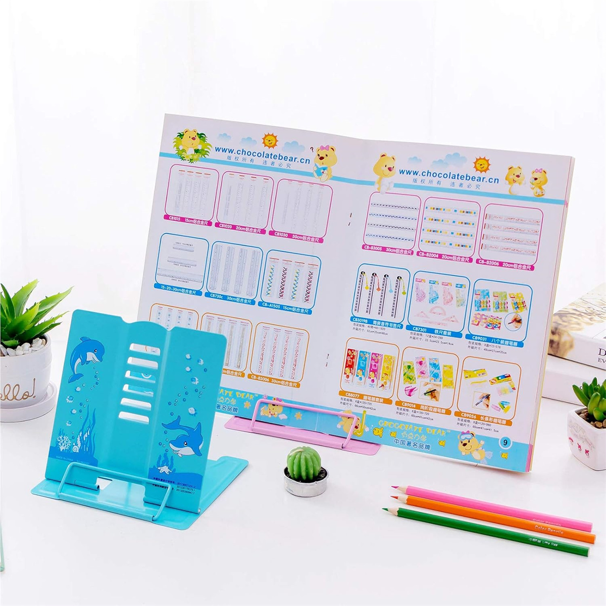 Ein blauer Buchständer mit Delfindesign hält einen offenen Katalog. Buntstifte und kleine Pflanzen stehen auf dem Tisch.