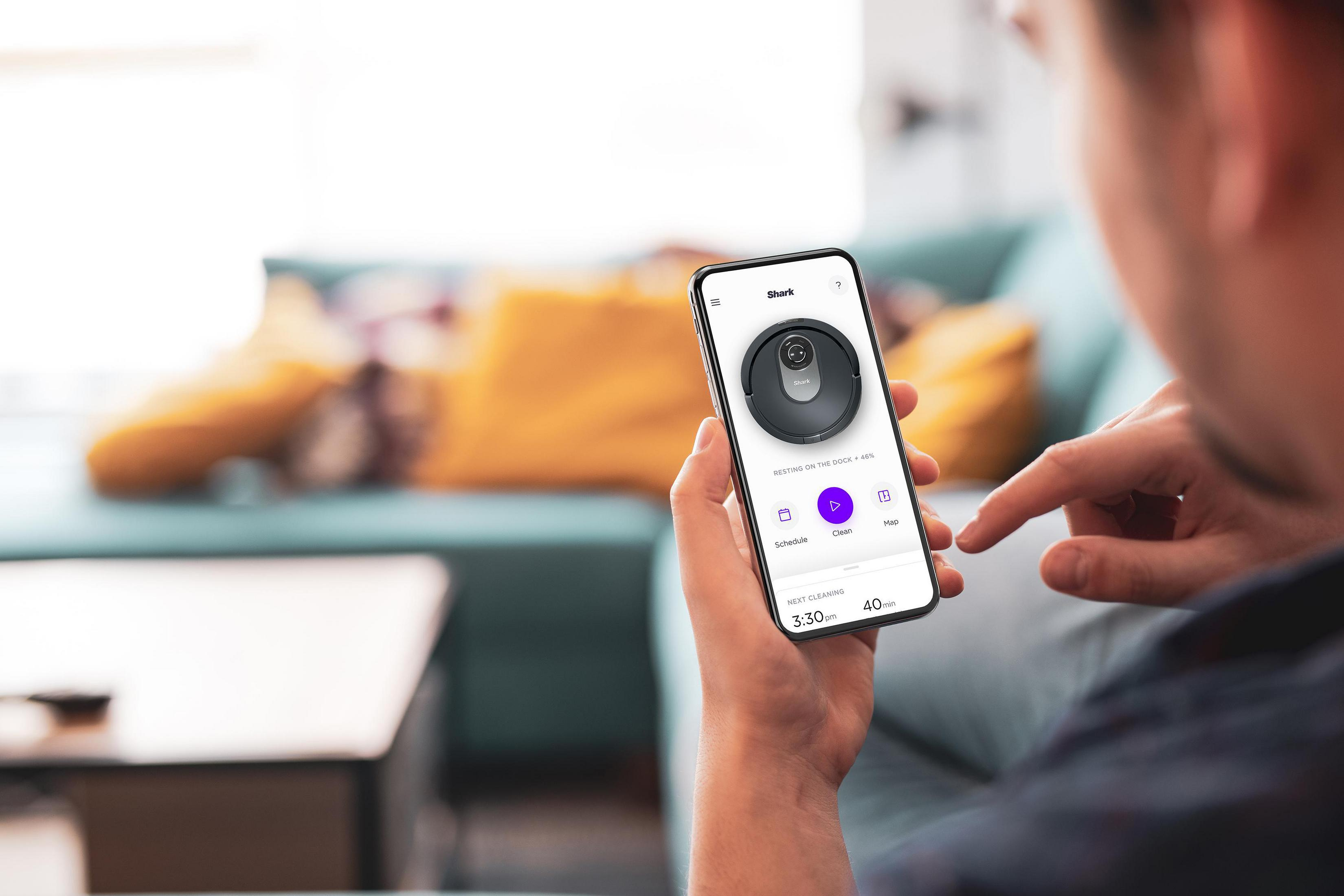 Eine Person hält ein Telefon, das eine Roboter-Staubsauger-App in einem Wohnzimmer zeigt.