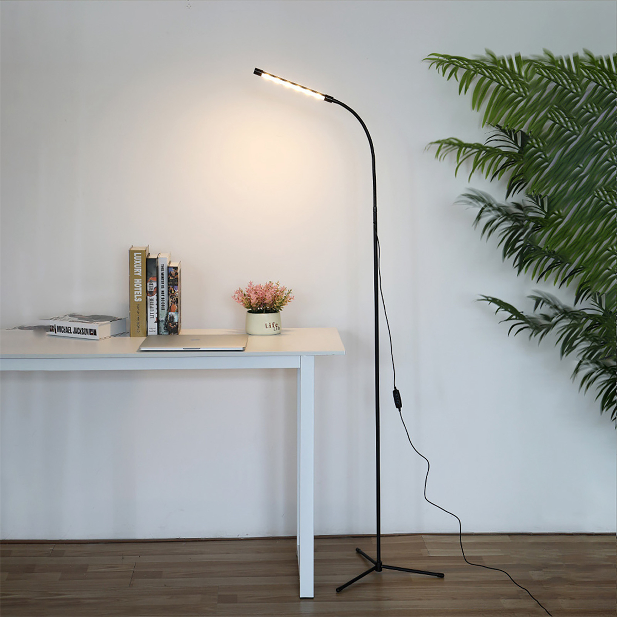 Eine hohe schwarze Stehlampe mit einem weißen Schreibtisch, Büchern und einer Pflanze. Das Licht ist an.