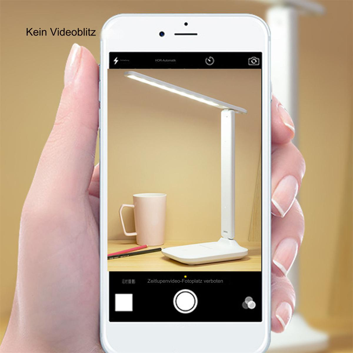 Eine Person macht mit einem Telefon ein Foto einer weißen Schreibtischlampe. Die Lampe ist an.