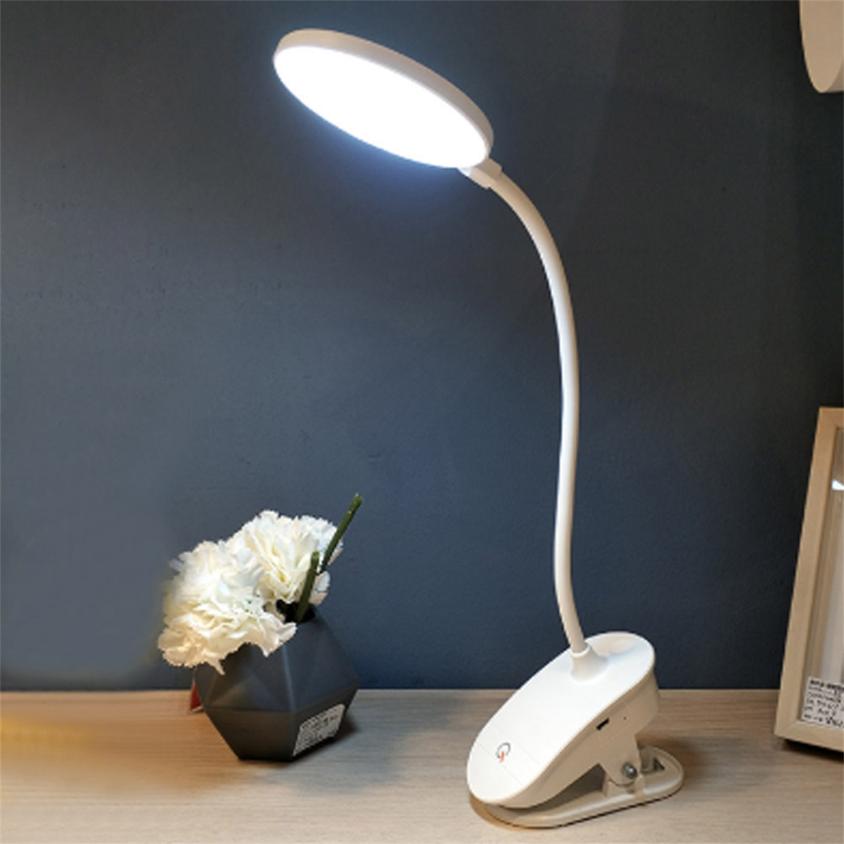 Weiße Klemm-Schreibtischlampe mit gebogenem Hals, Blumen und einem grauen Schreibtisch.