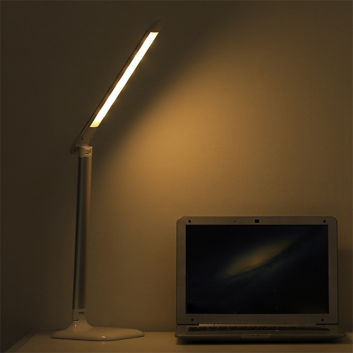 Eine weiße Schreibtischlampe mit einem beleuchteten Balken. Ein Laptop steht daneben. Der Hintergrund ist eine Wand.