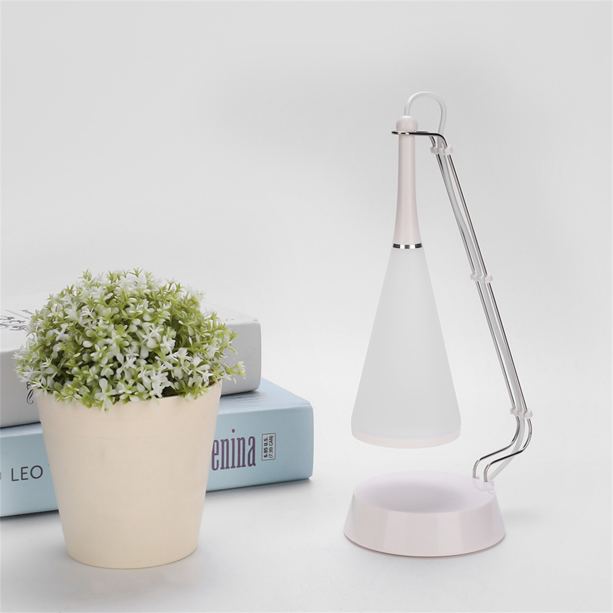 Weiße Schreibtischlampe mit konischem Schirm, neben einer Topfpflanze und Büchern.
