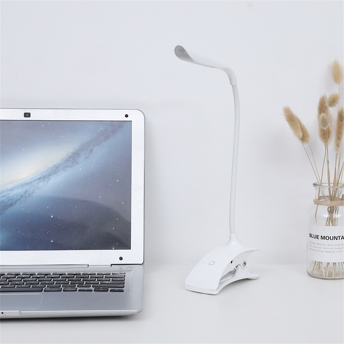 Eine weiße Klemm-Schreibtischlampe mit flexiblem Arm, neben einem Laptop und einer Blumenvase.