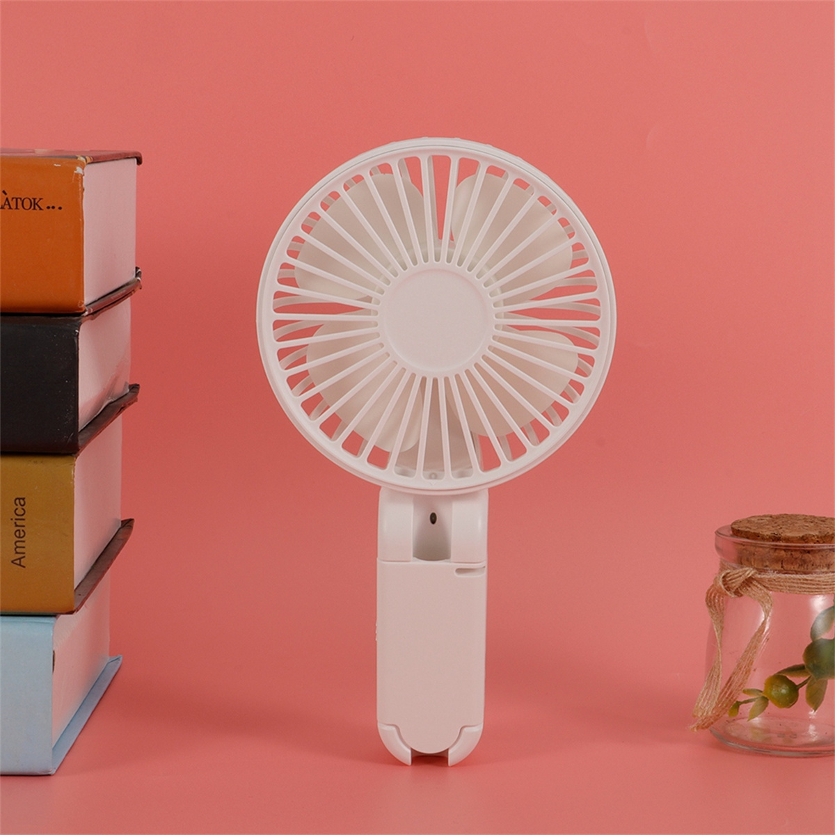 Ein weißer, tragbarer Ventilator mit weißen Flügeln, einem weißen Griff und rosa Hintergrund.