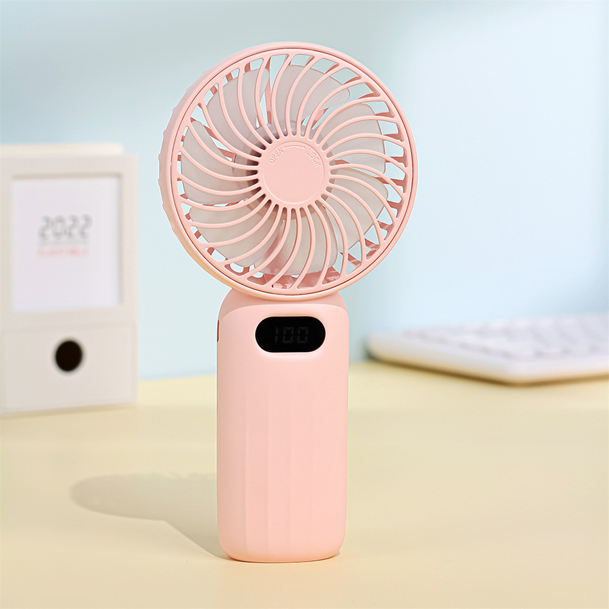 Rosa Handventilator mit Digitalanzeige, die '100' anzeigt. Oben befindet sich ein weißer Ventilator.