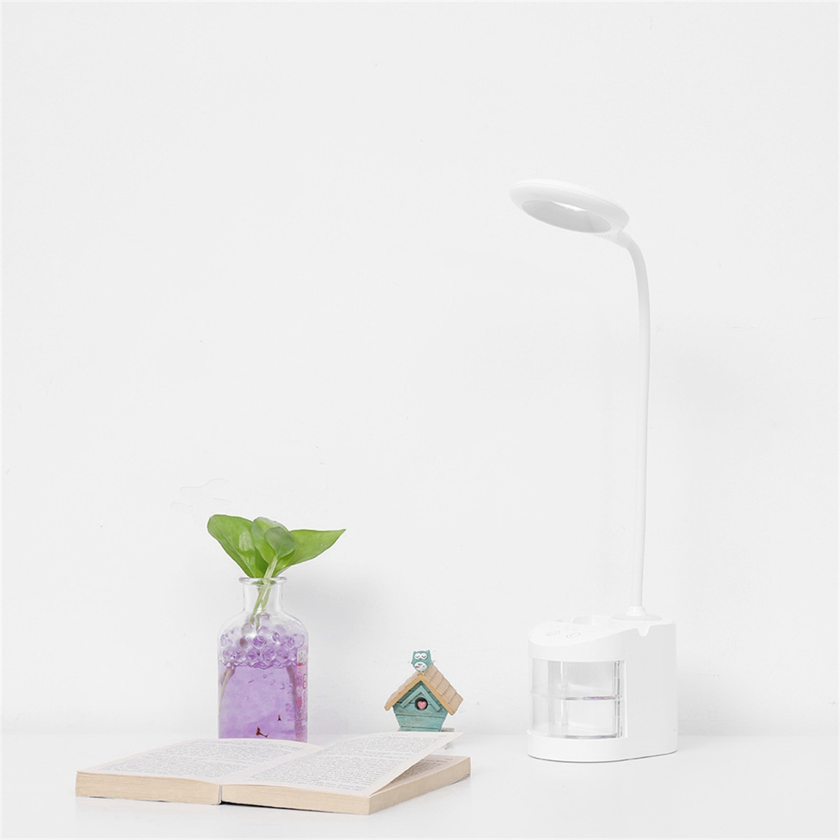 Weiße Schreibtischlampe mit gebogenem Licht. Eine kleine Vase mit einer Pflanze, ein offenes Buch und ein kleines Haus stehen auf dem Tisch.