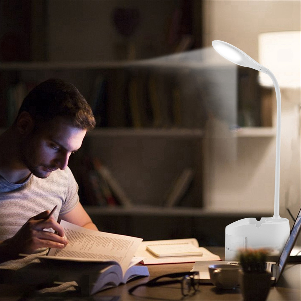 Ein Mann liest ein Buch unter einer weißen Schreibtischlampe. Bücher und eine Tasse sind in der Nähe.