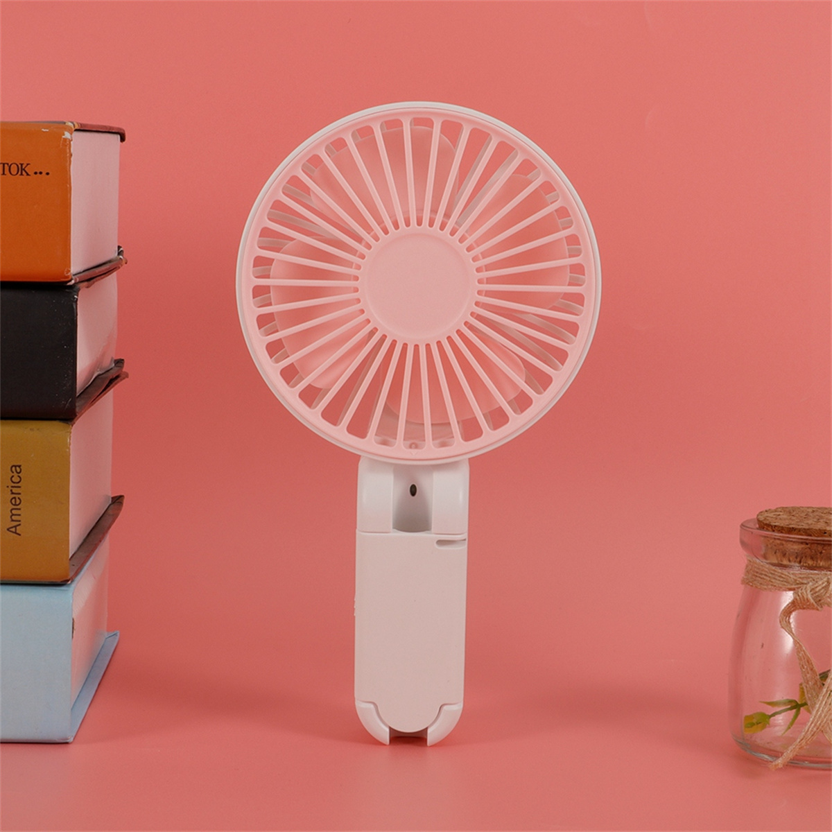 Weiß-rosa tragbarer Ventilator ist geöffnet, auf rosa Oberfläche mit Büchern und einem Glas.