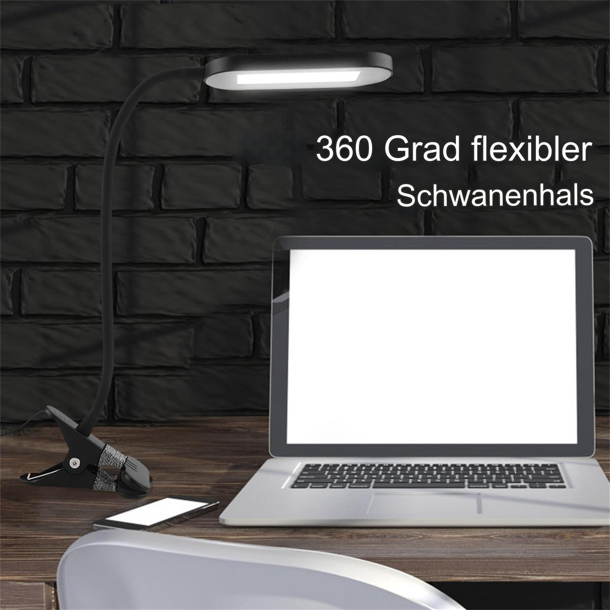 Eine schwarze Schreibtischlampe an einem Schreibtisch befestigt, mit Laptop und Telefon. Die Lampe ist an.