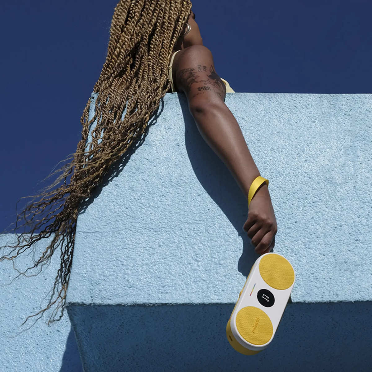 Kobieta z długimi włosami i tatuażem, trzymająca żółto-biały głośnik. Niebo jest niebieskie.