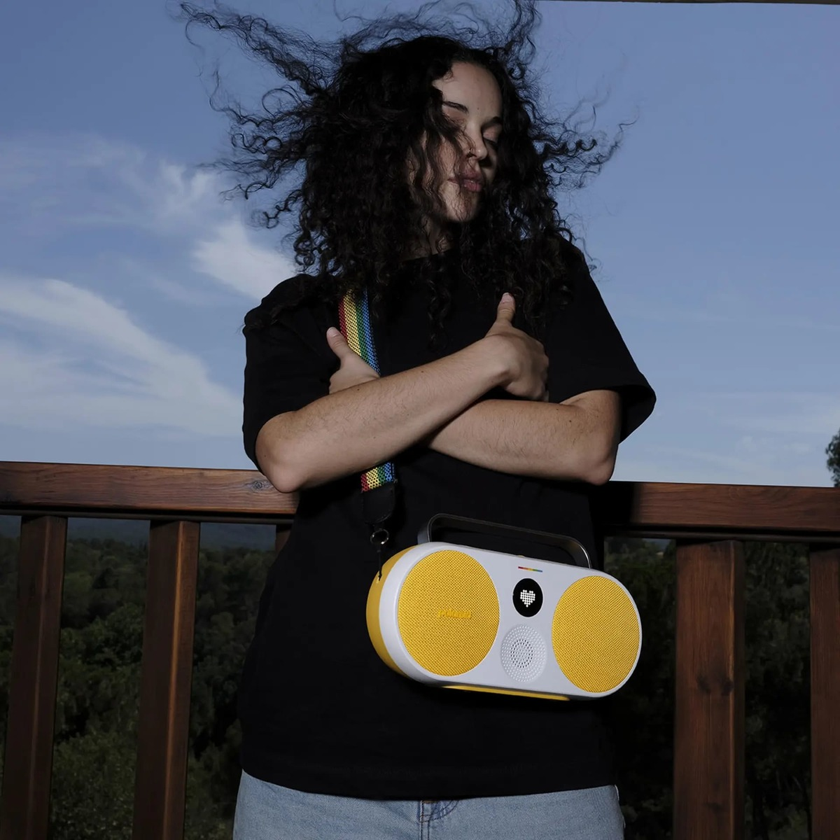Osoba z kręconymi włosami stoi z żółto-białym przenośnym głośnikiem na ramieniu, na zewnątrz.
