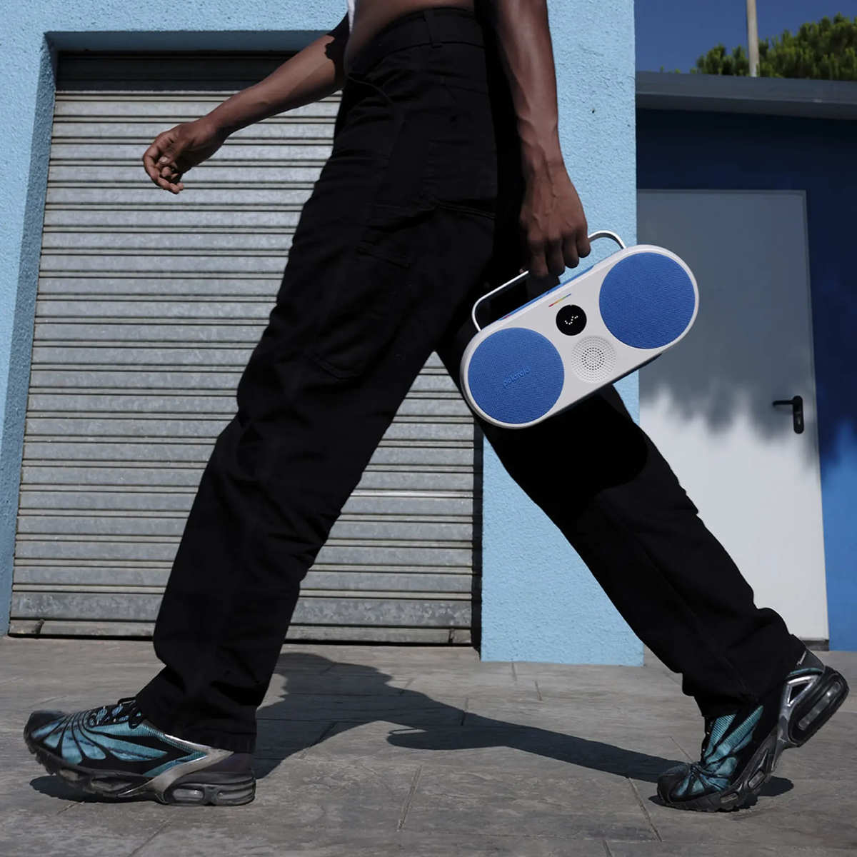 Osoba idzie z niebiesko-białym głośnikiem. Nosi czarne spodnie i buty, tło niebieskiej ściany.