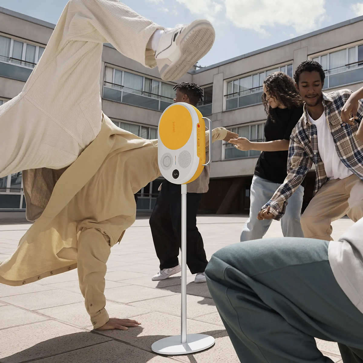Ludzie tańczą wokół żółto-białego głośnika na srebrnym słupie. Jedna osoba tańczy breakdance.