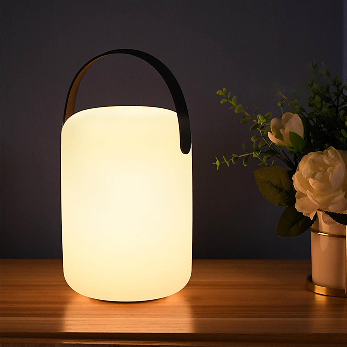 Weiße Lampe mit Griff, leuchtend, auf Holztisch, Pflanzen daneben.