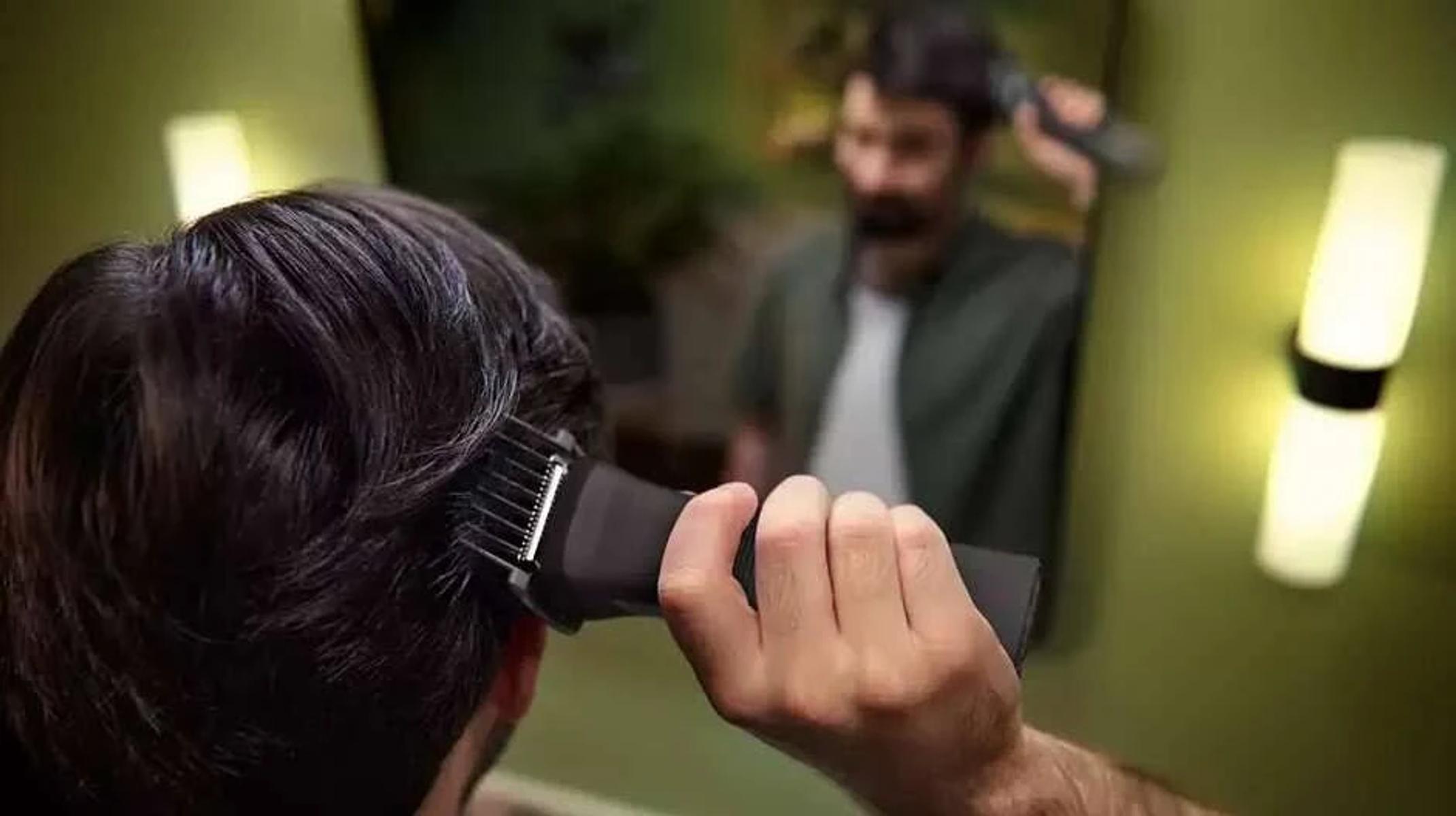 Un hombre se corta el pelo con una máquina en un espejo. El fondo está borroso.