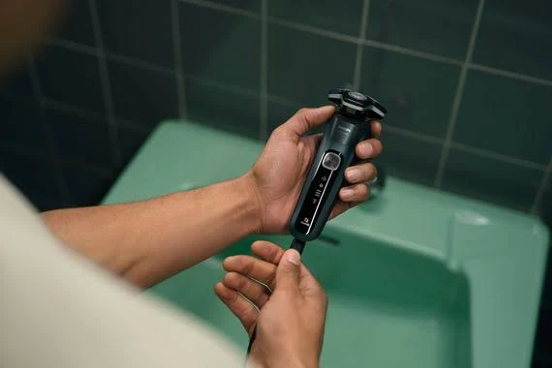 Mains d'un homme tenant un rasoir électrique noir, près d'un lavabo vert clair.