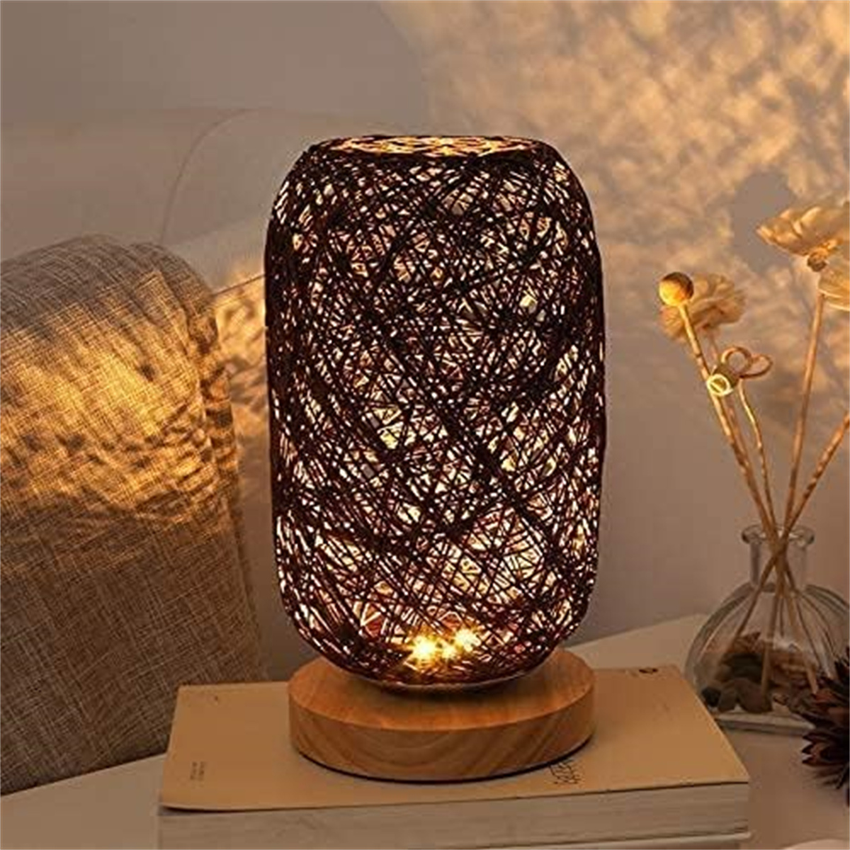 Eine braune, gewebte Lampe auf einem Holzsockel, die auf einem Buch steht. Ein graues Kissen und Blumen im Hintergrund.