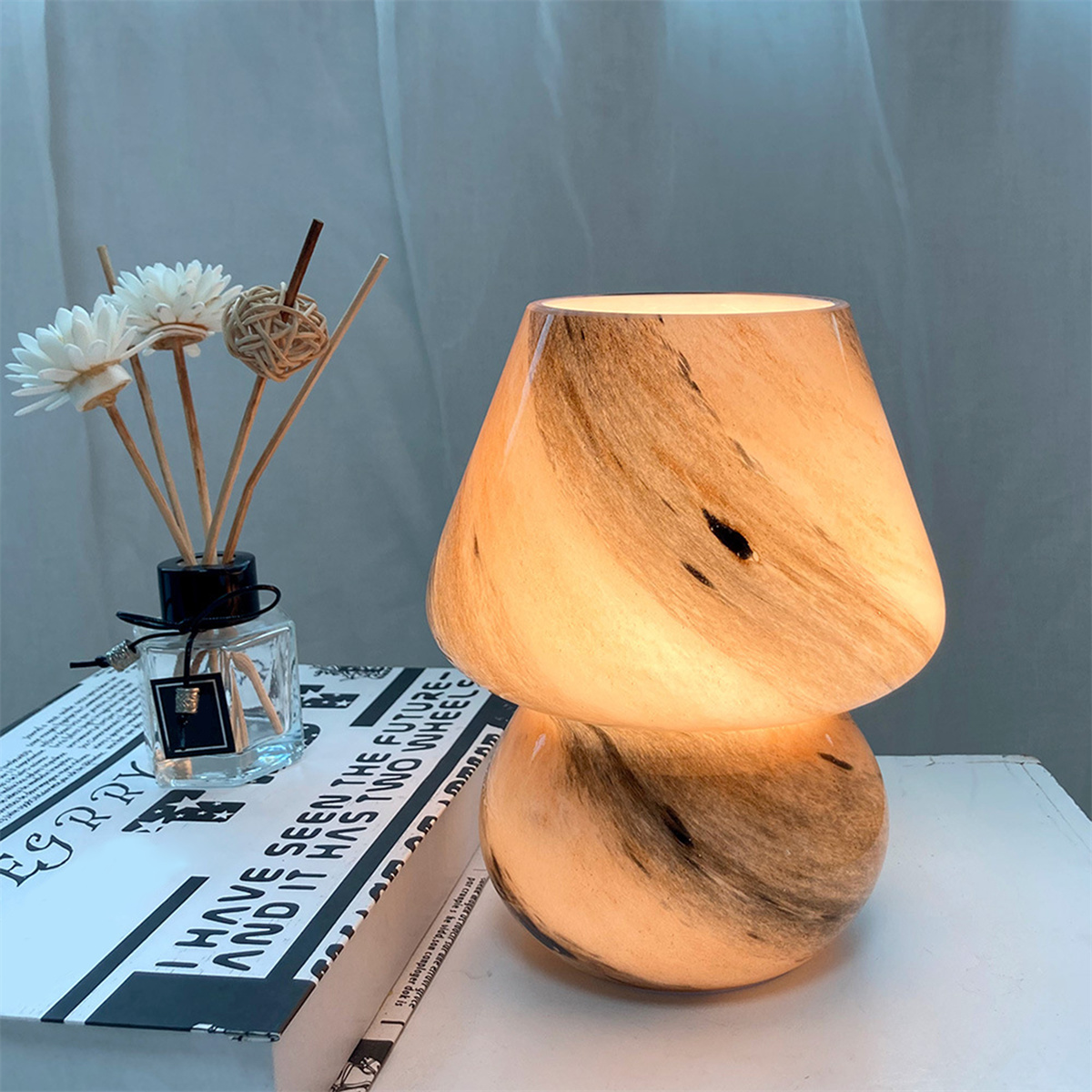 Eine pilzförmige Lampe mit Marmormuster, neben einem Diffusor mit Blumen und einem Buch.