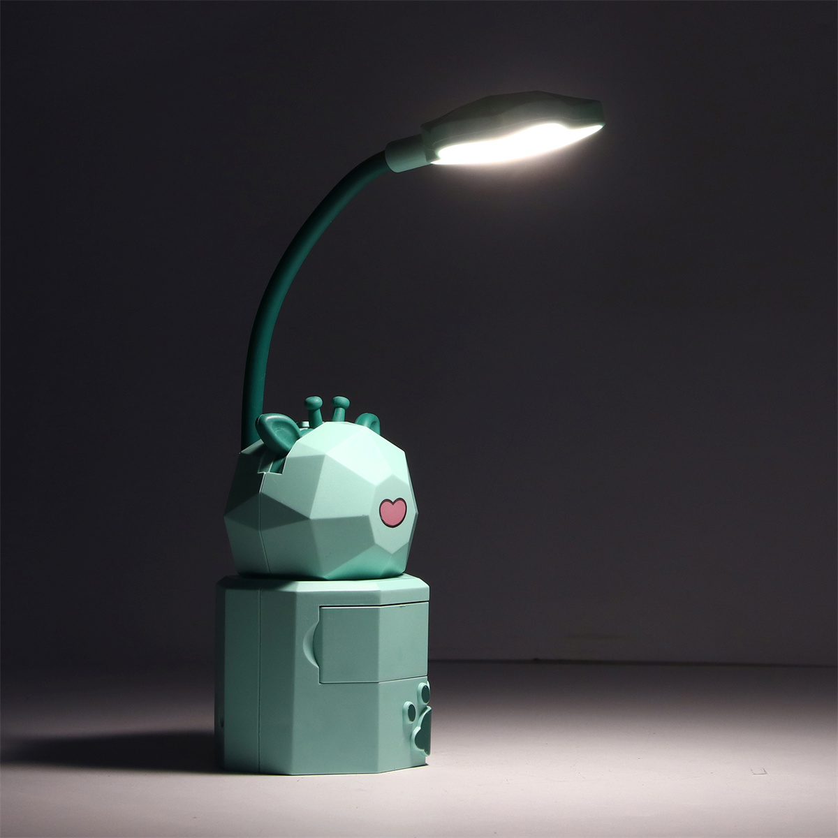 Eine grüne Lampe in Giraffenform. Die Lampe steht auf einem würfelförmigen Sockel. Der Hintergrund ist dunkel.
