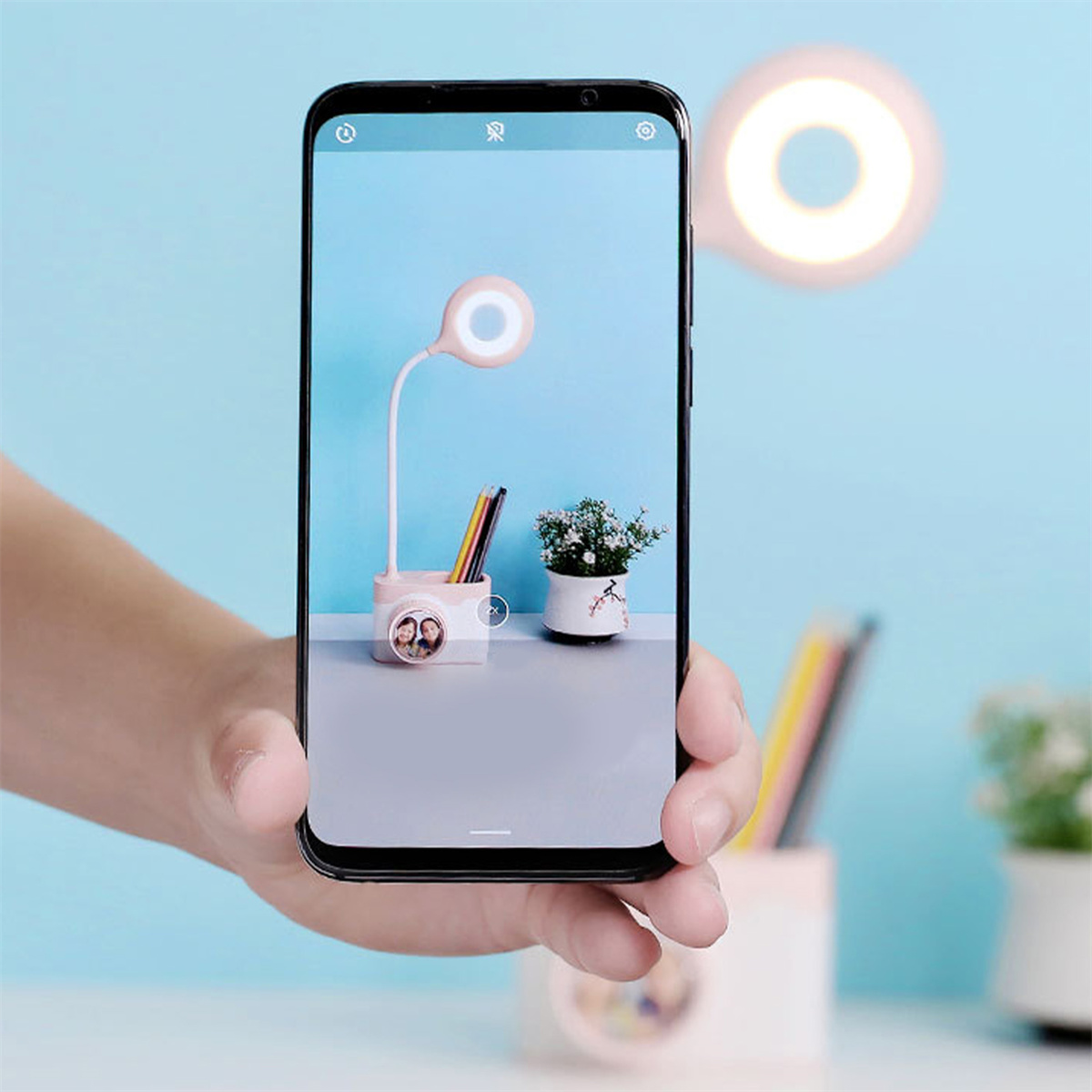 Ein Telefon, das eine Schreibtischlampe mit Ringlicht, Bleistiften und einer kleinen Pflanze anzeigt. Das Telefon wird von einer Hand gehalten.