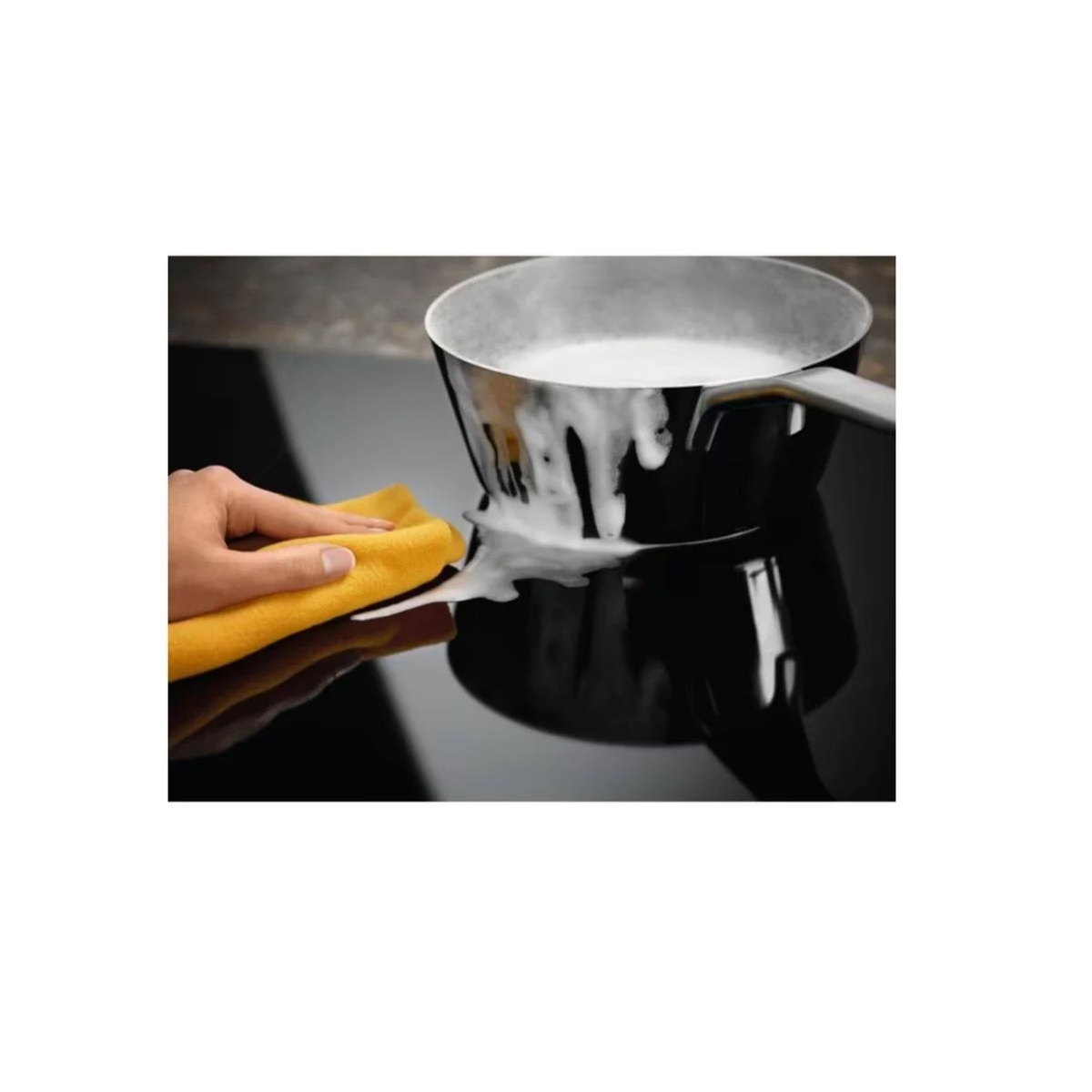 Ręka wyciera czarny garnek żółtą szmatką. Biały płyn wylewa się z garnka.