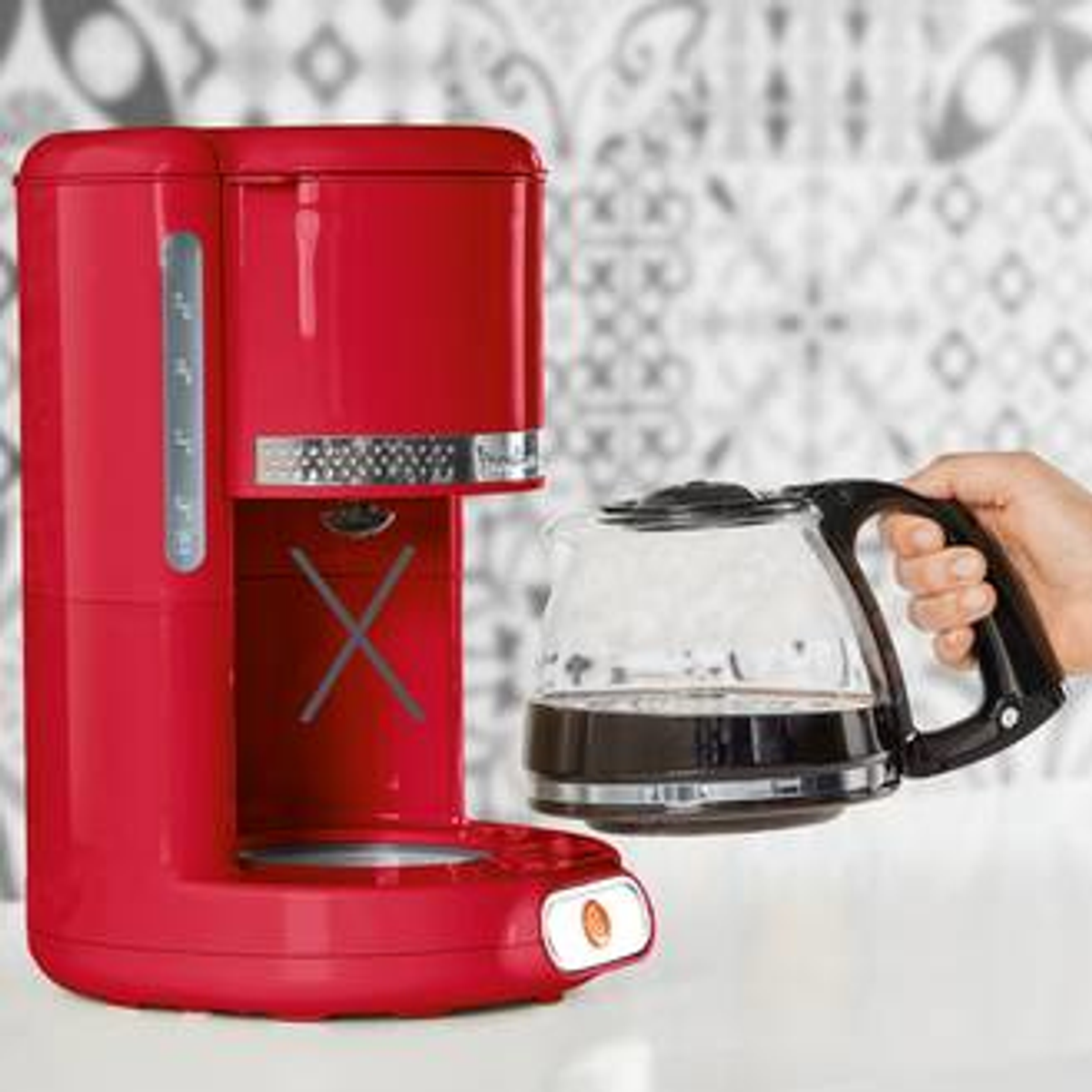 Czerwony ekspres do kawy ze szklanym dzbankiem trzymanym przez rękę na wzorzystej powierzchni.