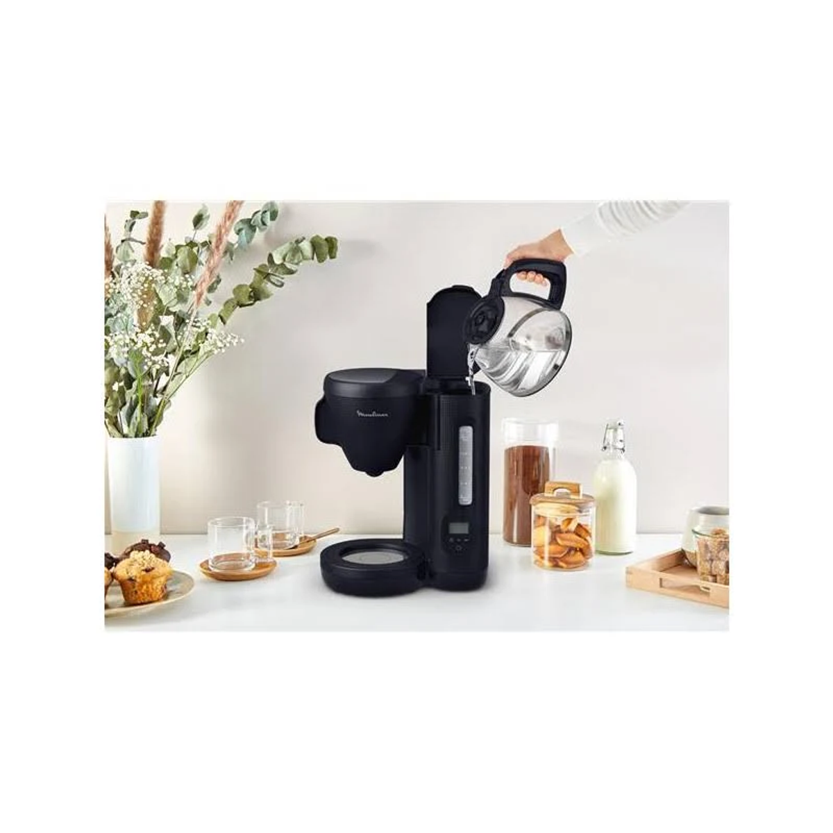 Black coffee maker with water being poured into it. White background with muffins and jars.