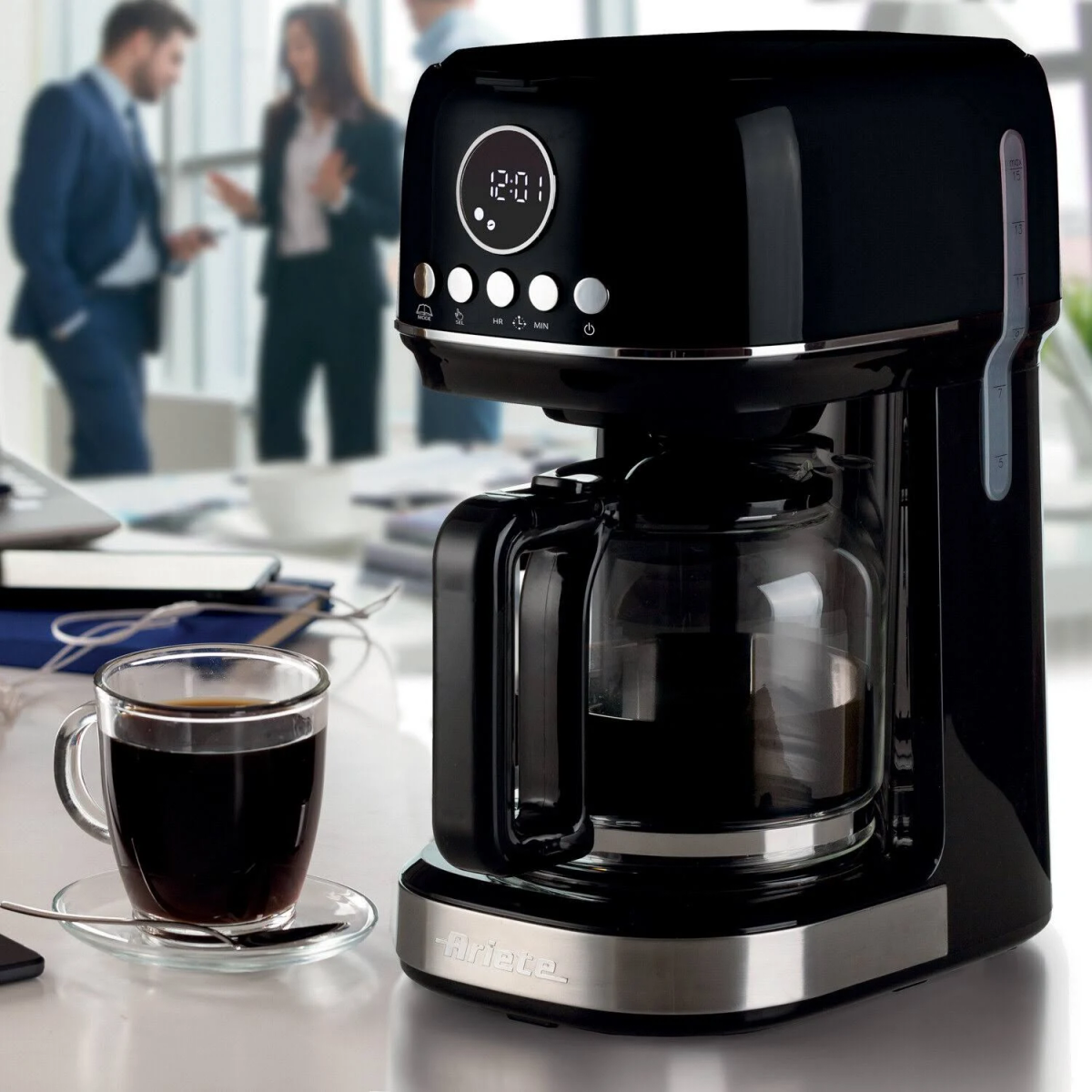 Czarny ekspres do kawy ze szklaną karafką. Filiżanka kawy na spodku, ludzie w tle.