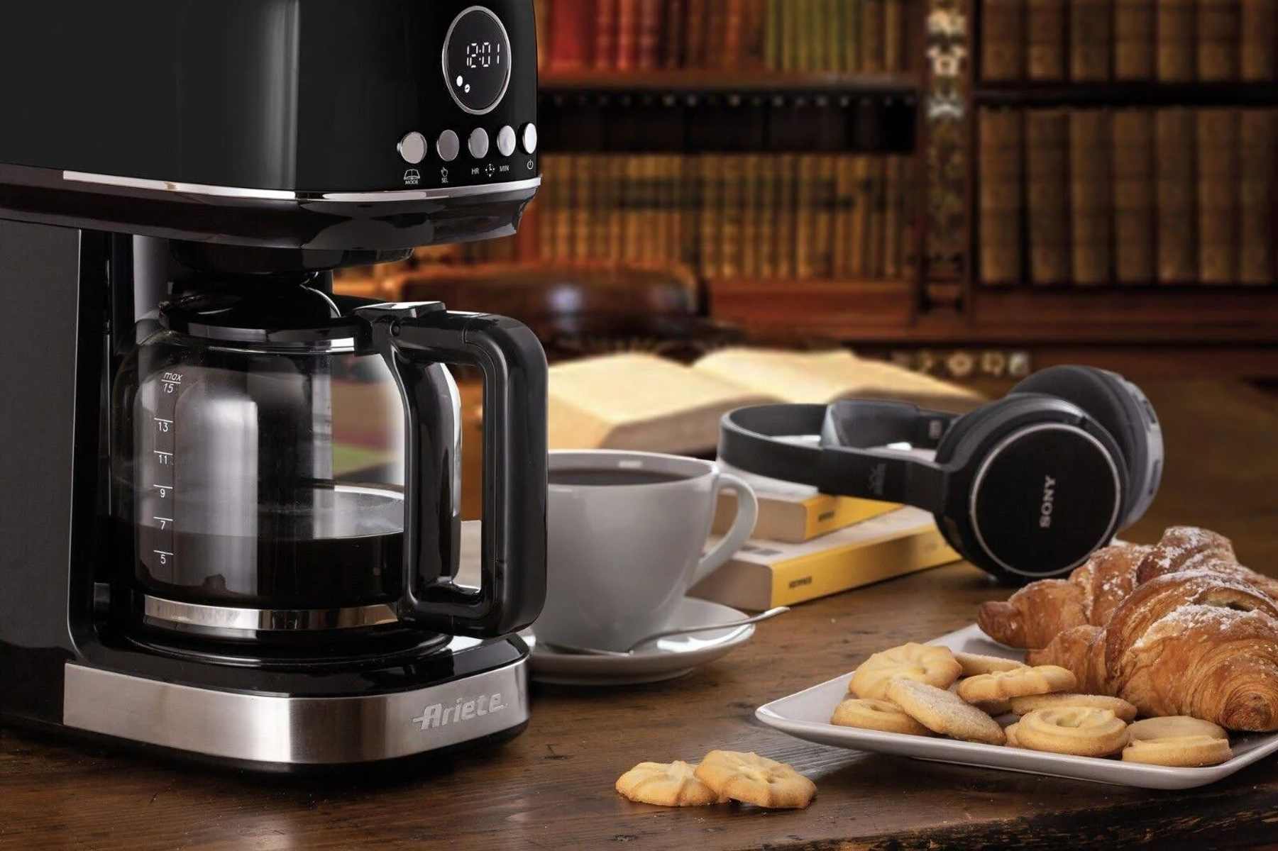 Ekspres do kawy, filiżanka kawy, ciasteczka, rogaliki i słuchawki na drewnianym stole.